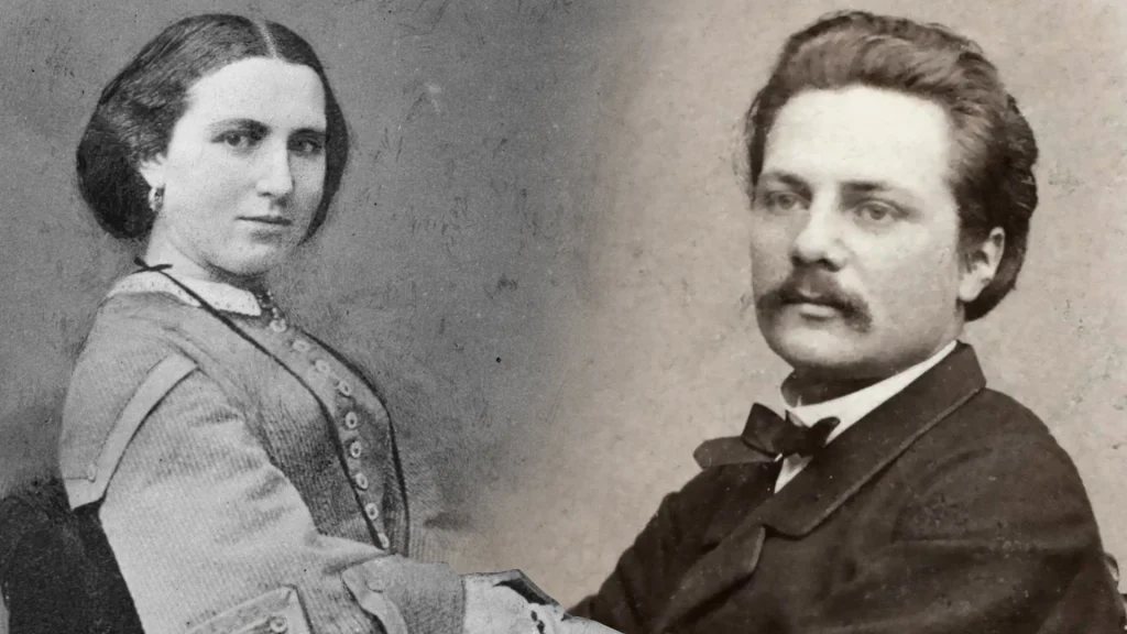 A young couple in the 19th century, the woman with dark hair styled up, wearing a light dress, a long necklace, and earrings, gazes into the camera. The man to the right, dressed in a suit and bowtie, sporting a mustache with slicked-back hair and crossed arms, directs his gaze outside the frame.