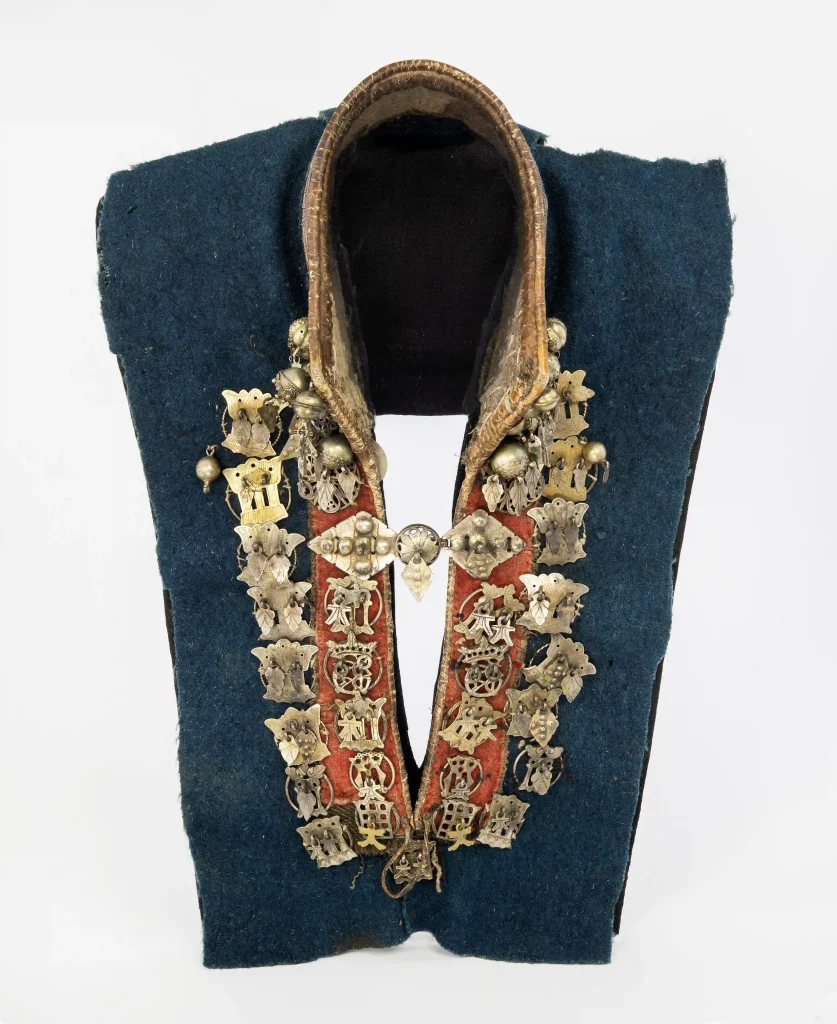 Krage som sitter på ett blått tyg med röd kant och guldfärgade broderier, framtill helt klädd med silverhängen i olika former