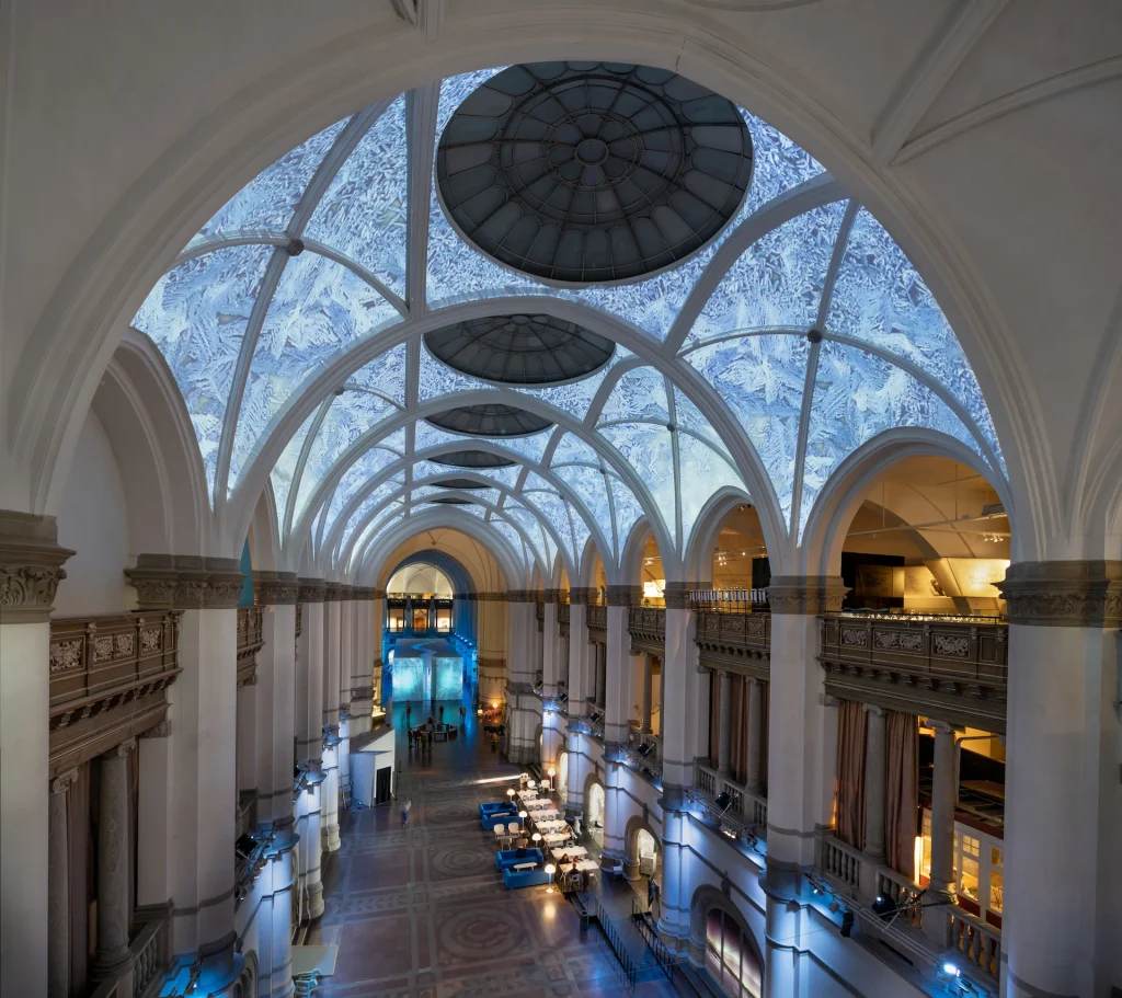 Nordiska museets stora hall i helbild med projektioner i takvalven som föreställer rimfrost, längst bort syns ingången till utställningen Arktis i form av ett stort isblock