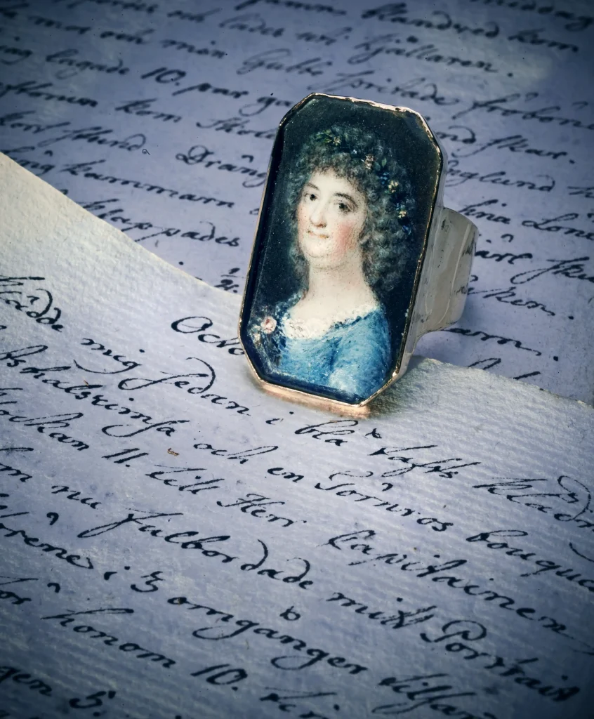 En ring med ett porträtt av en 1700-talsdam ligger på dagbokssidor med snirklig handstil skriven med gåspenna.