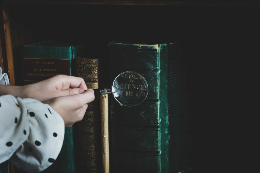 Barnhänder håller ett förstoringsglas mot tjocka gamla bokryggar i en bokhylla.