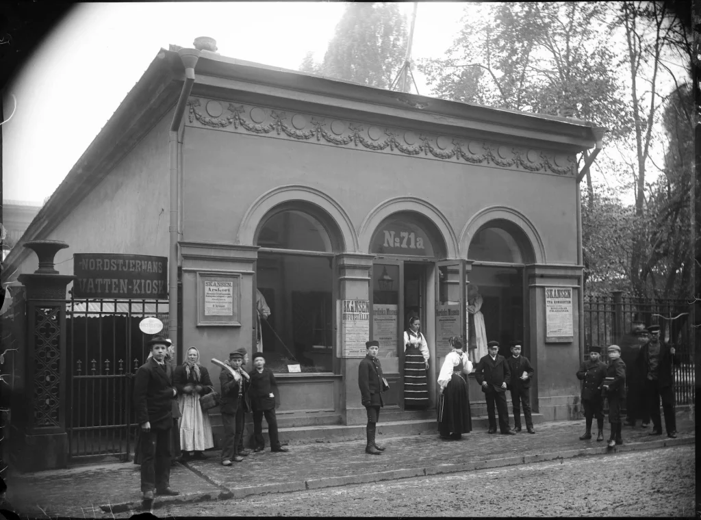 A stone house with three arched windows and children and adults at the entrance looking towards the camera. A 'dalkulla' in traditional costume stands in the doorway.