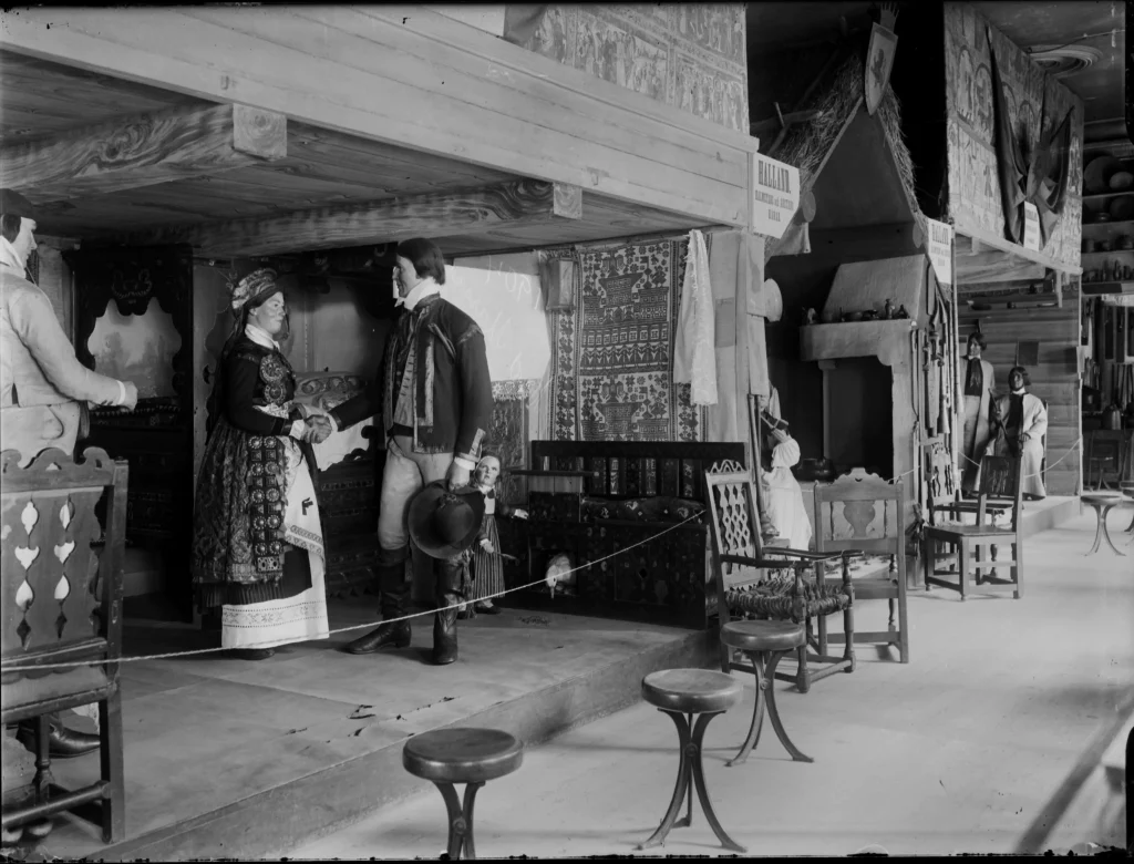 A hall with a series of constructed rooms. The closest one resembles a cottage with dolls representing a woman and two men in traditional costumes. A small girl doll is visible behind the man.