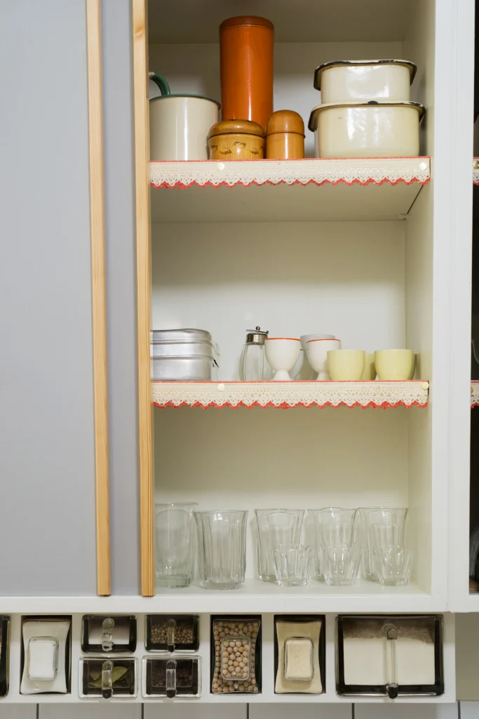 Ett köksskåp med skjutlucka och trälist visar glas och koppar på en hylla med spetskant.