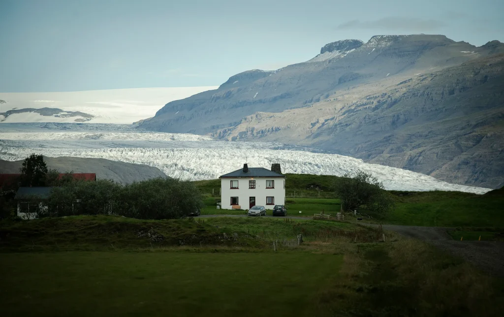 Naturvy från Island med ett vitt bostadshus i mitten på grön barmark, i bakgrunden frusen havsvik och högt berg