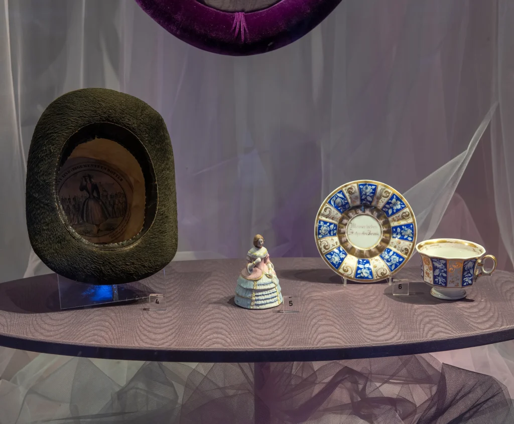 Föremål i museimonter: porslinsfigurin, en hatt och kaffekopp.