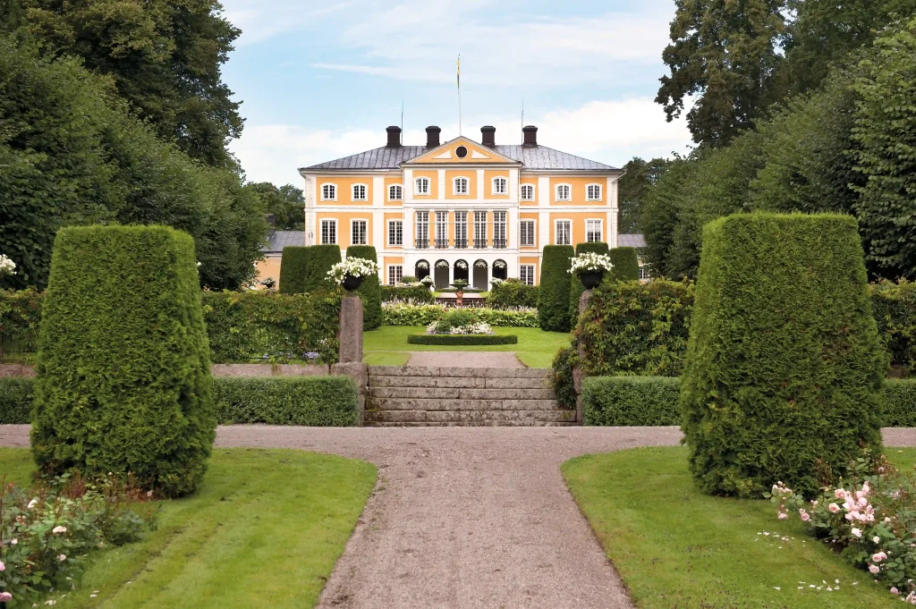 "Large yellow manor house with well-trimmed bushes and a gravel path in the foreground."