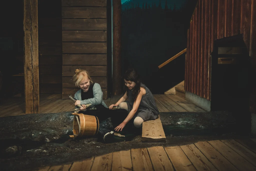 Två flickor skrattar och fyller en spann med vatten vid en låtsas-å.