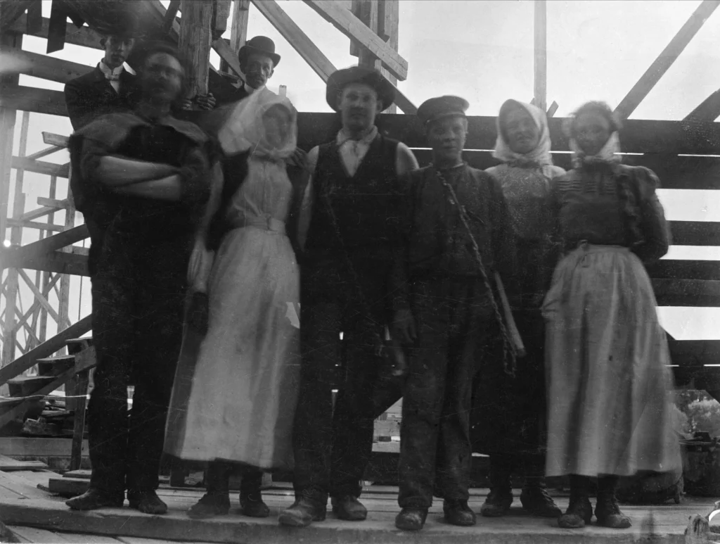 Eight people stand on a scaffolding, including men, women, and young boys.