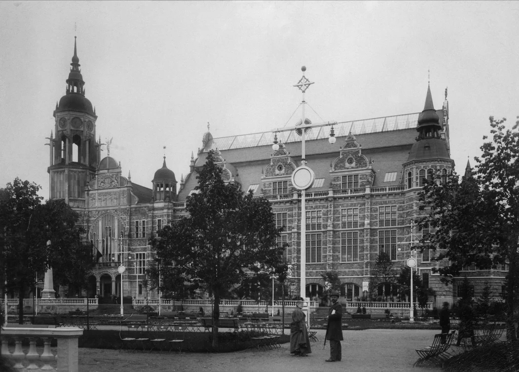A partially constructed building and people in 19th-century attire among park benches and plantings on Djurgårdsvägen.
