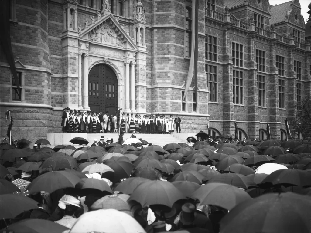 At the entrance to a grand castle-like building, Dalecarlian women in traditional costumes stand on the stairs. Below the stairs, a crowd with umbrellas gathers.