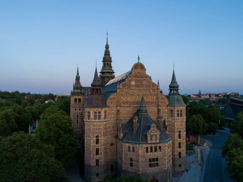 Stor byggnad med tinnar och torn i skymningsljus. Fasadbokstäver med texten Nordiska museet.