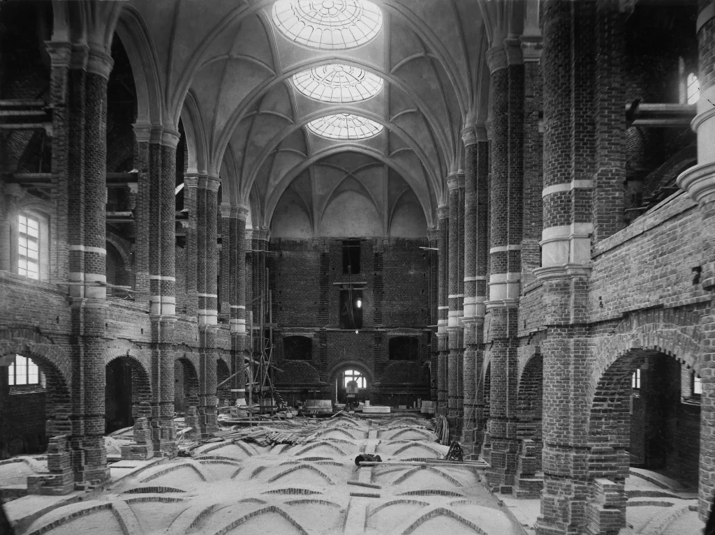 A large hall with arches, columns, and skylights that is not yet completed. Raw brick and an unfinished floor are visible.