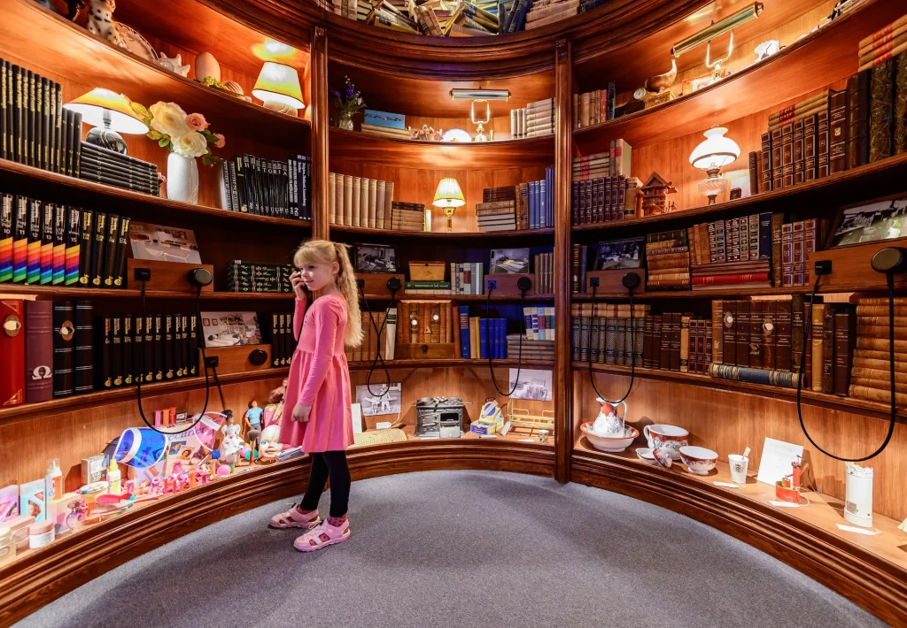 En flicka står och lyssnar i hörlurar vid en bokhylla med många detaljer och föremål