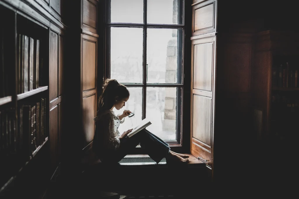 En flicka sitter i ett fönster och tittar i en bok, hon håller ett förstoringsglas i handen.