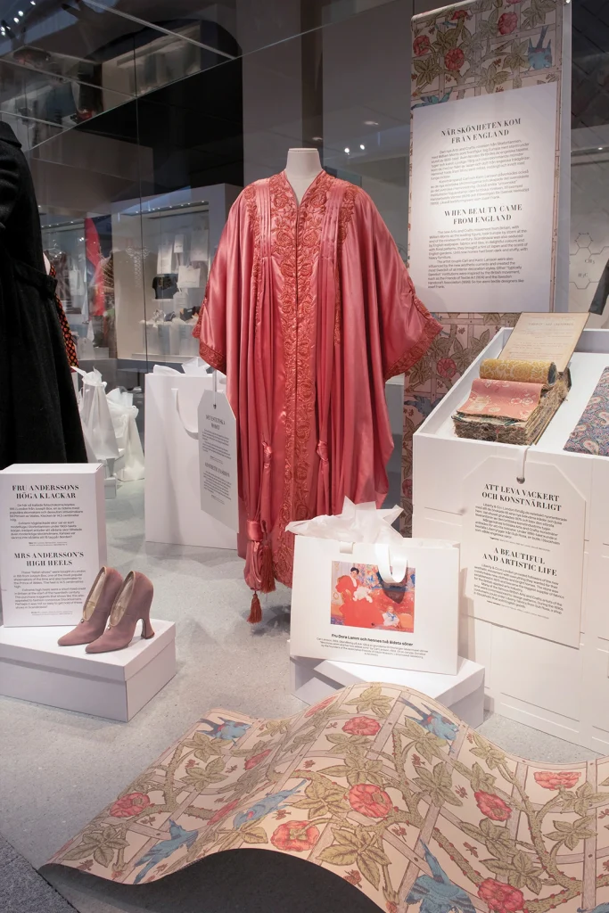 En monter visar högklackade skor och en rosa kaftan, nederst i bild syns ett engelskt tapetmönster från Arts & Crafts