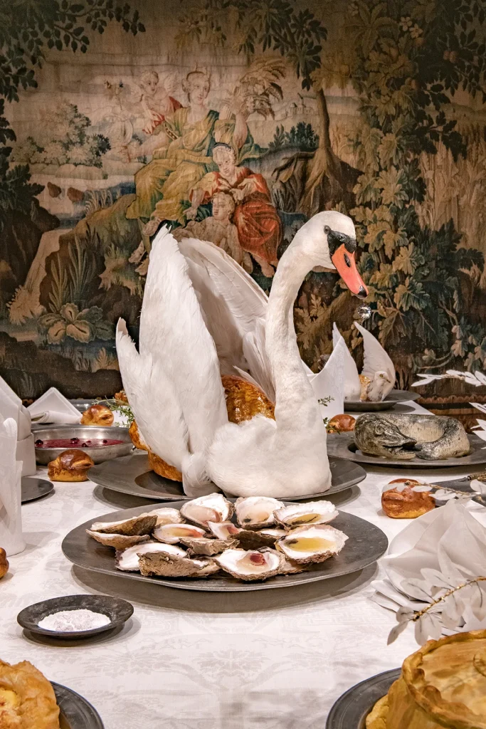 Ett bord är dukat för fest med tenntallrikar och ostron, mitt på bordet en dekoration i form av en uppstoppad svan med en stek i.