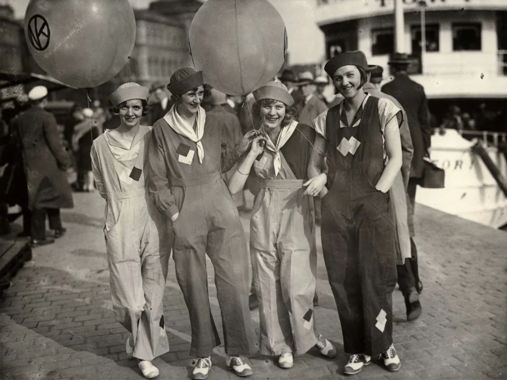 Fyra kvinnor klädda i overaller. promenadskor, scarfar runt halsen och uppvikta tygsolmössor ler och håller i reklamballonger.