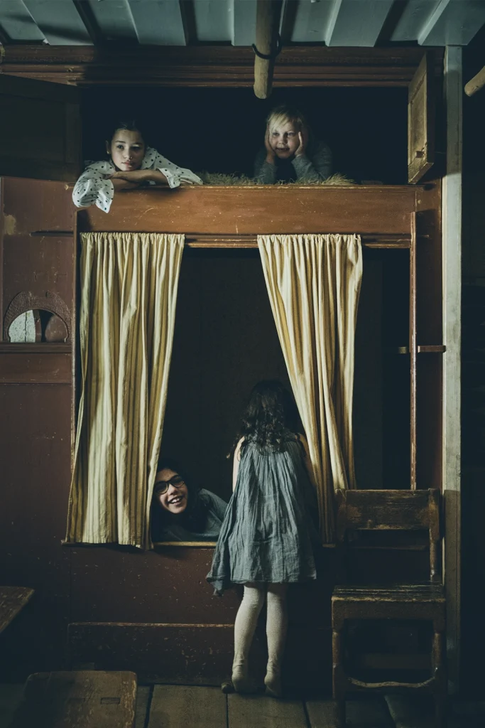 En sovalkov med förhängen och överslaf, fyra barn i olika åldrar har krupit upp i sängarna på och leker.