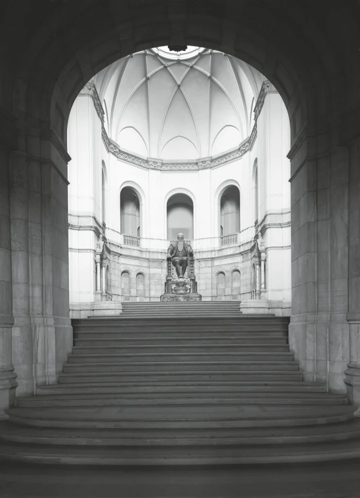 En trappa leder upp till en stor sal med takfönster, valv och pelare mot en stor staty av en äldre Gustav Vasa i gips.