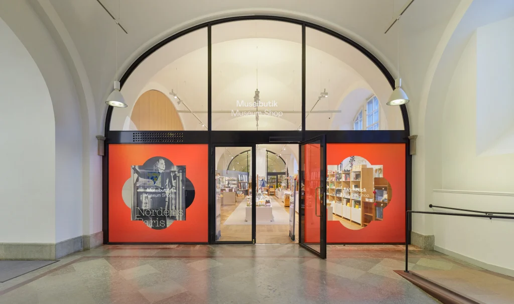 En ljus entréhall med höga valv och glasad ingång till en museibutik. Glasväggarna har en orange inramning i form av Nordiska museets symbol.
