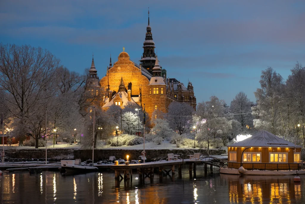 Nordiska museet sett från vattnet en vinterdag i skymningen, ett varmt gult sken lyser mot fasaden.