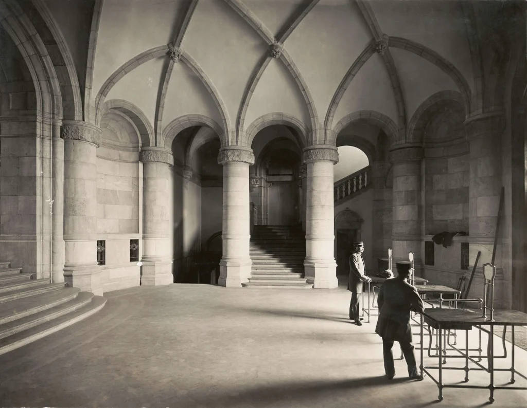 In a large hall with vaults and pillars, two museum guards in uniform stand by separate gates, looking out towards an entrance outside the frame. Daylight streams in.