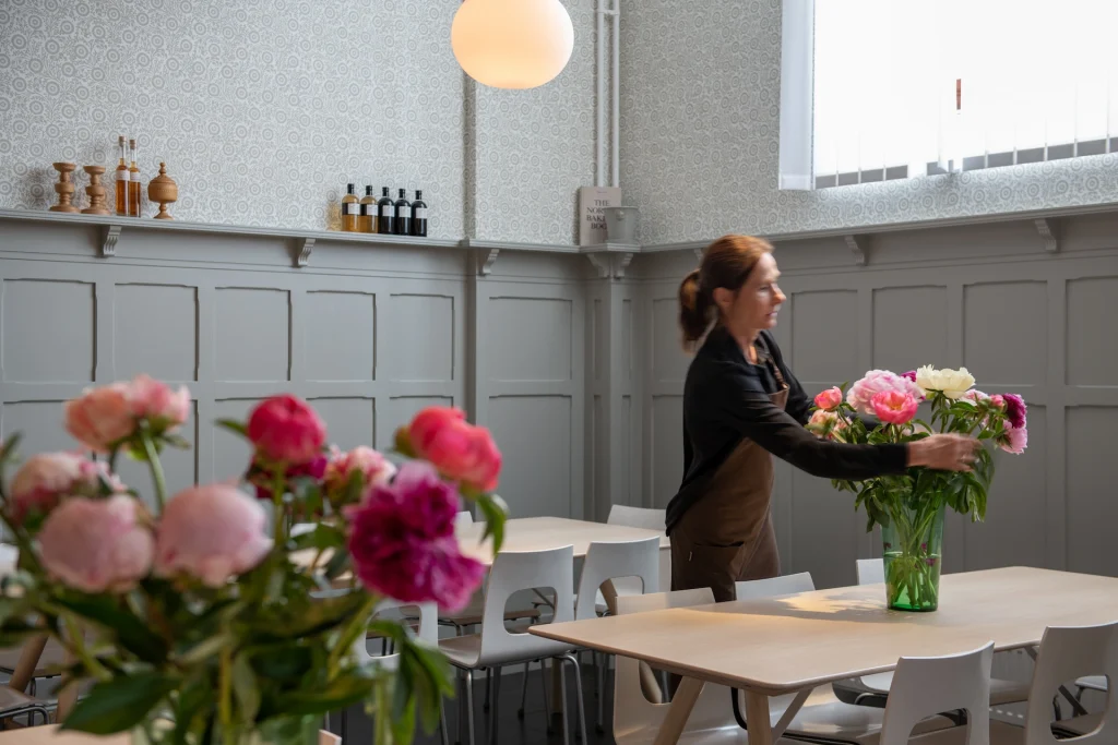 En kvinna arrangerar en vas med pioner i en restaurang med träpanel och mönstrade tapeter i ljus grågrönt.