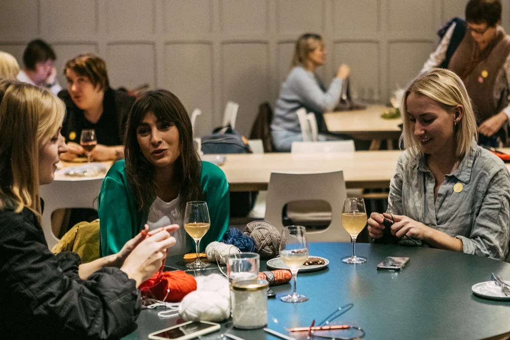 Tre unga kvinnor sitter i en restaurang och stickar och dricker ett glas vin.