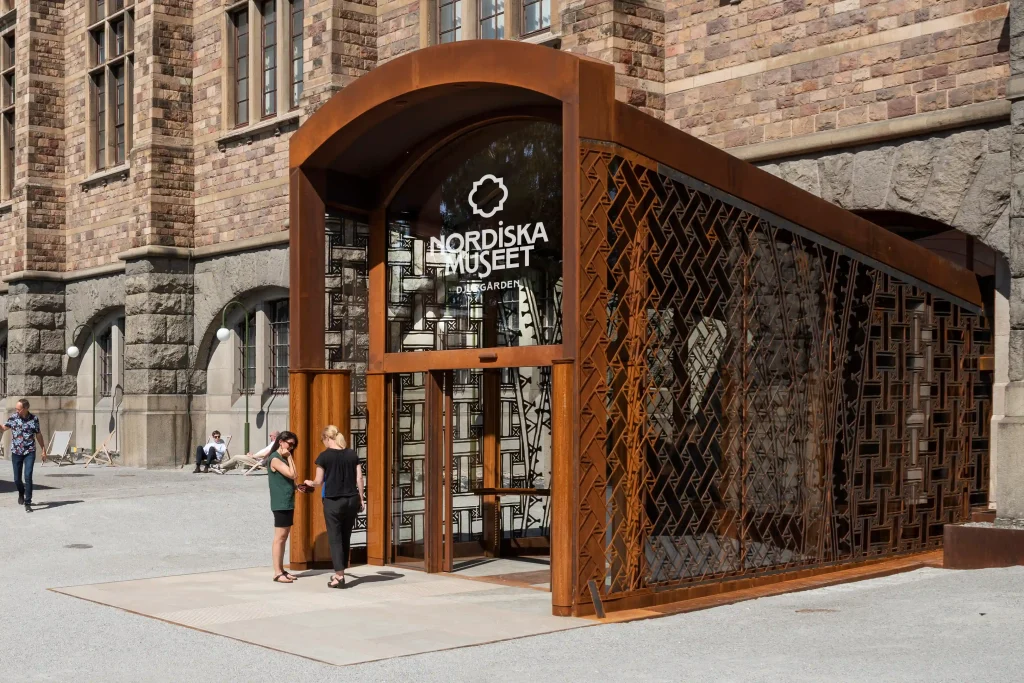 A protruding portal in corten steel with glass walls featuring a pattern. Two women stand at the entrance.