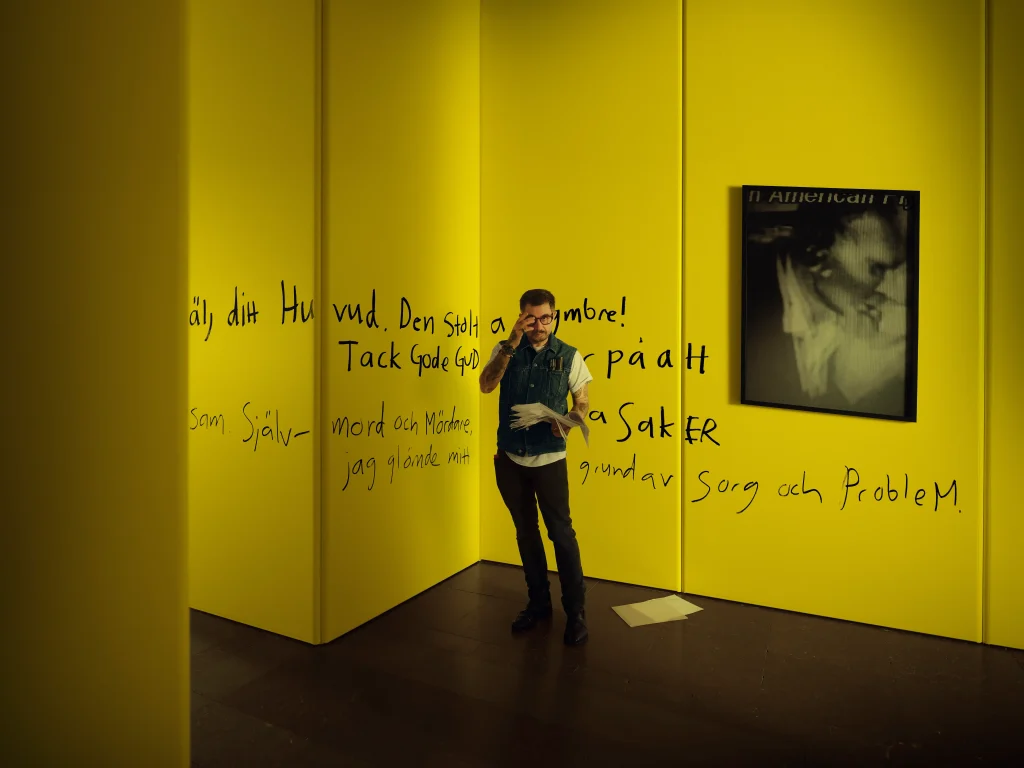 En man i jeansväst och glasögon står i ett gult rum vid ett inramat fotografi, på väggen är ord skrivna med tjock svart tuschpenna.