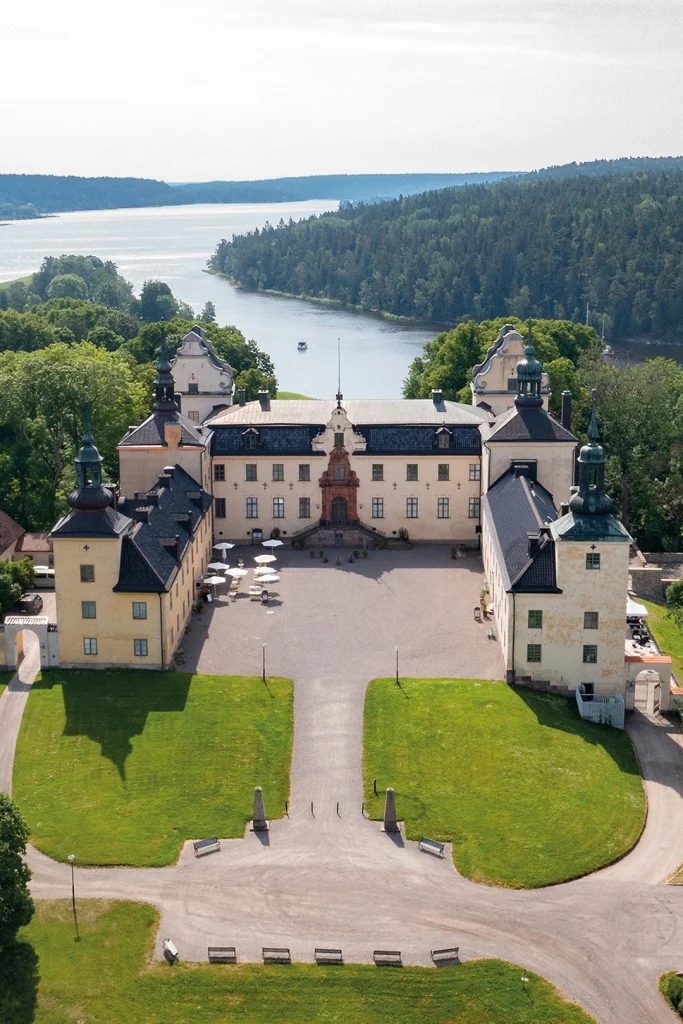 Ett slott och borggård med lummiga träd, stora gräsytor och glittrande fjärd i bakgrunden.