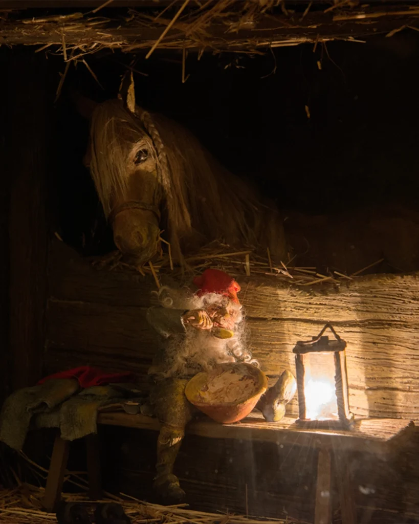 Ett tittskåp med ett stall med häst där en hustomte sitter och äter gröt vid en liten tänd lykta.