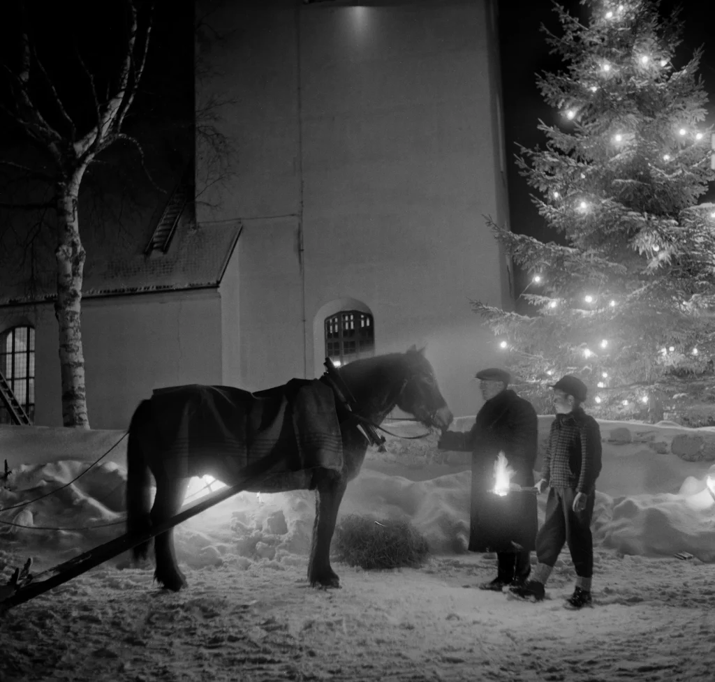 En man och en ung pojke håller grimman på en häst med släde utanför en kyrka en snöig julotta, pojken håller i en fackla och en julgran syns bakom dem.