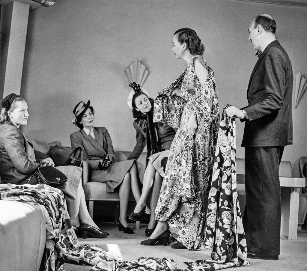 En mannekäng visar kläder för tre damer som sitter i en soffa, en man står bakom mannekängen och håller upp tyger.
