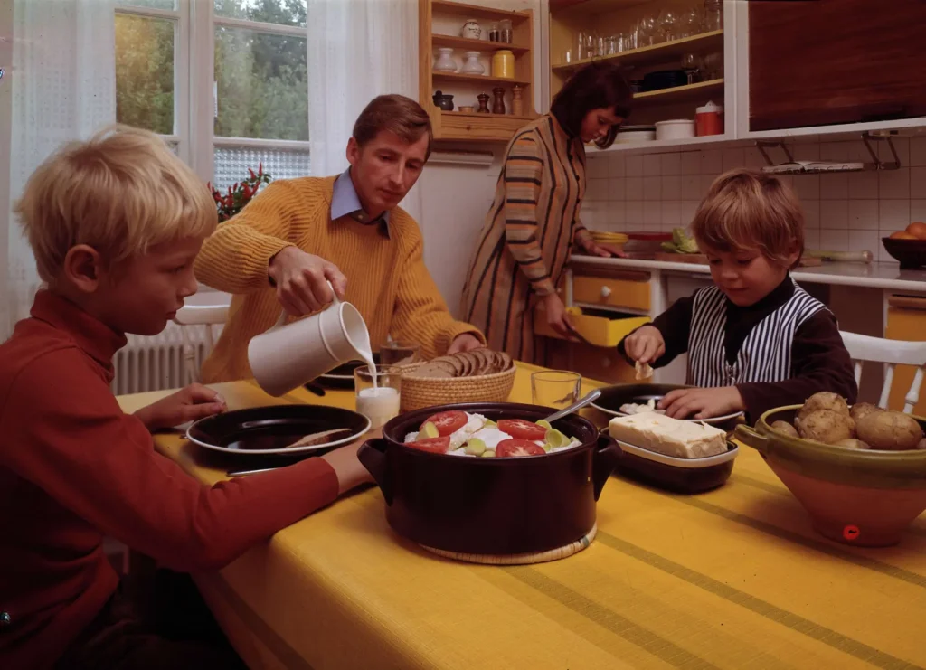 En familj vid ett köksbord i ett 70-talskök i gult och brunt, mamma drar ut en kökslåda, pappa häller upp mjölk till en pojke, den andra pojken brer en smörgås.