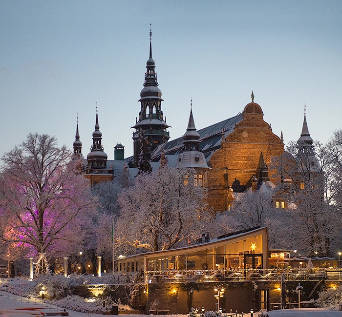 Nordiska museet i snö och skymning mot en ljus vinterhimmel. Det lyser ett varmt gult sken från fönstren.