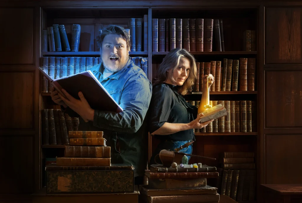 En man och en kvinna i ett mörkt gammalt bibliotek håller i en bok som det lyser ett magiskt sken ur, kvinnan håller i en pendel.