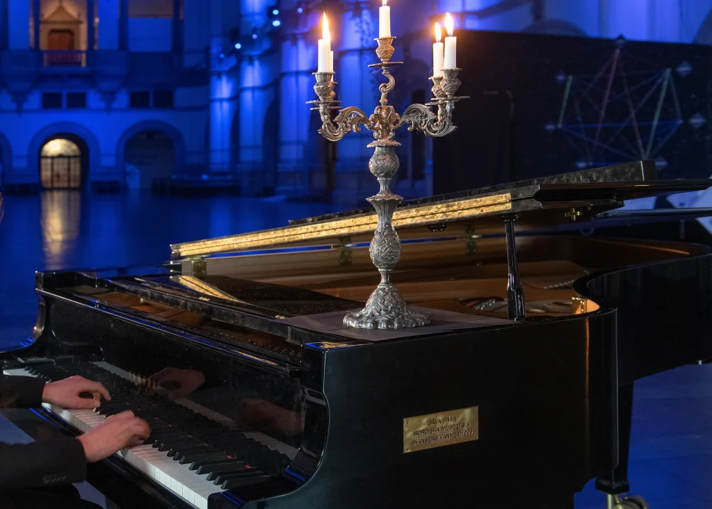 En flygel med tända ljus i en ljusstake, pianistens händer syns vid tangenterna, flygeln står i Nordiska museets stora hall som är ljussatt i ett blått sken.
