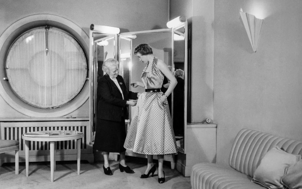En äldre dam poch en ung kvinna i sidenklänning står vid en spegel i ett exklusivt provrum och provar klänningen.