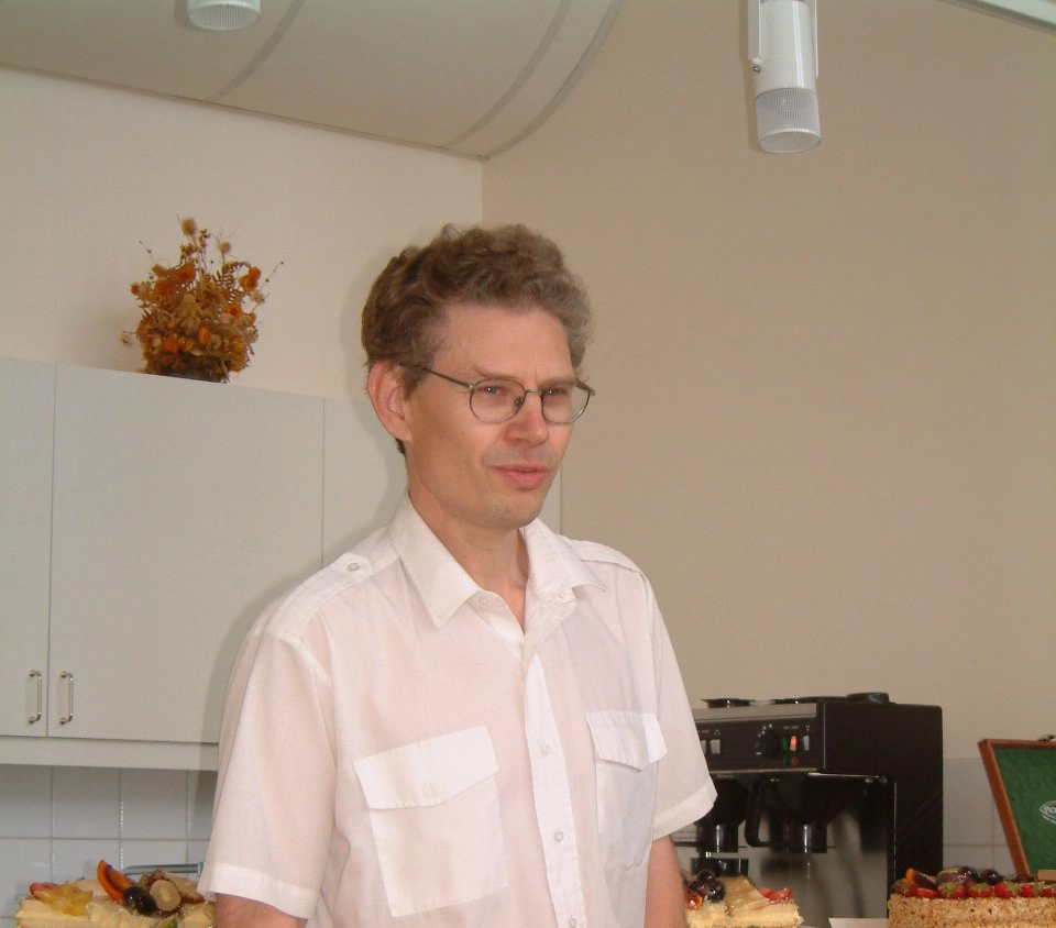 En man i 70-årsåldern med glasögon och vit kortärmad skjorta i ett kök. Bkom honom är tårtor och kaffe uppdukat.