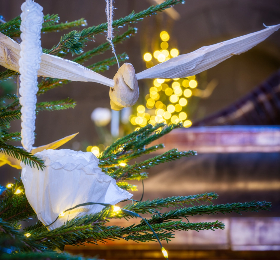 Närbild på en täljd träfågel och dekorationer i en julgran, i oskarp bakgrund anas fler granar och föremål.