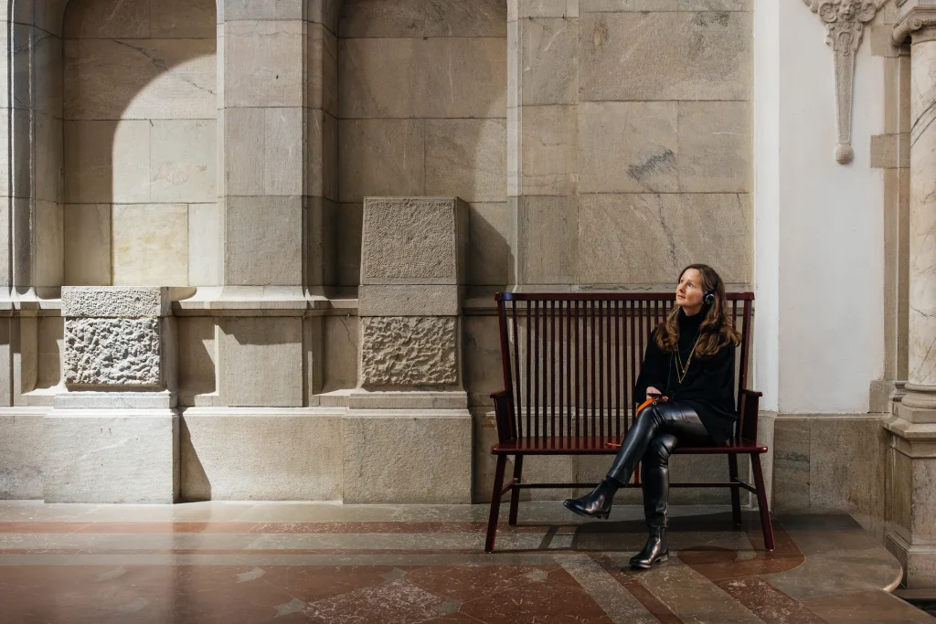 En kvinna med långt hår och hörlurar sitter på en vinröd pinnsoffa och lyssnar på audioguide i Nordiska museet, bakom henne marmorväggar med nischer och reliefer. Det lyser ett sken från det hon tittar mot när hon lyssnar.