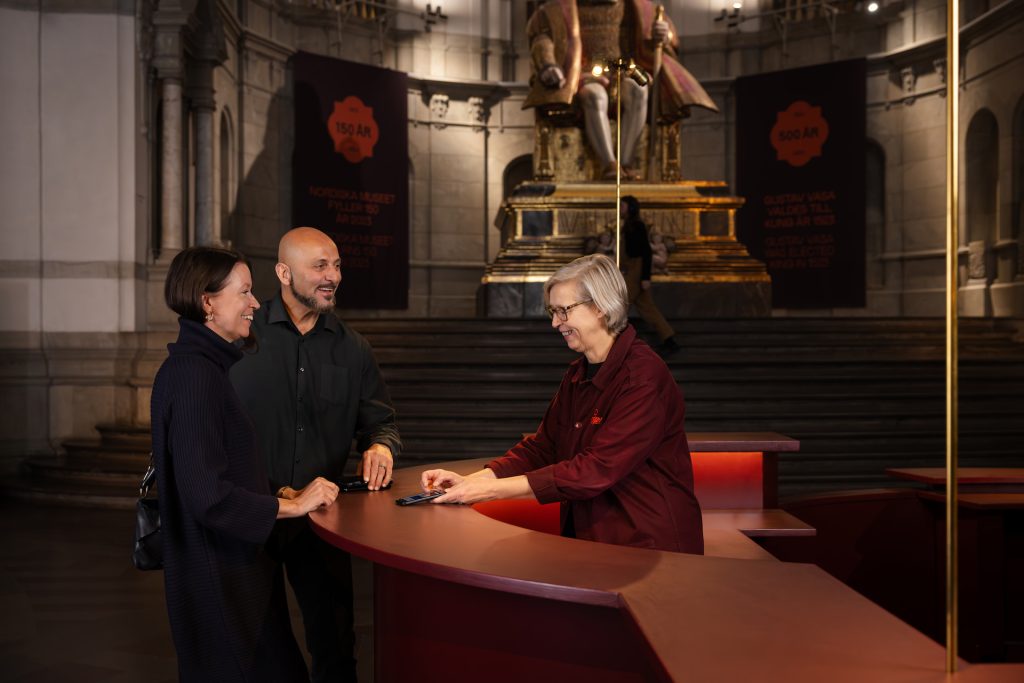 En värd i vinröd rock i Nordiska museets reception visar en man och en kvinna museets audioguide. Bakom skymtar den stora Gustav Vasa statyn och en besökare.