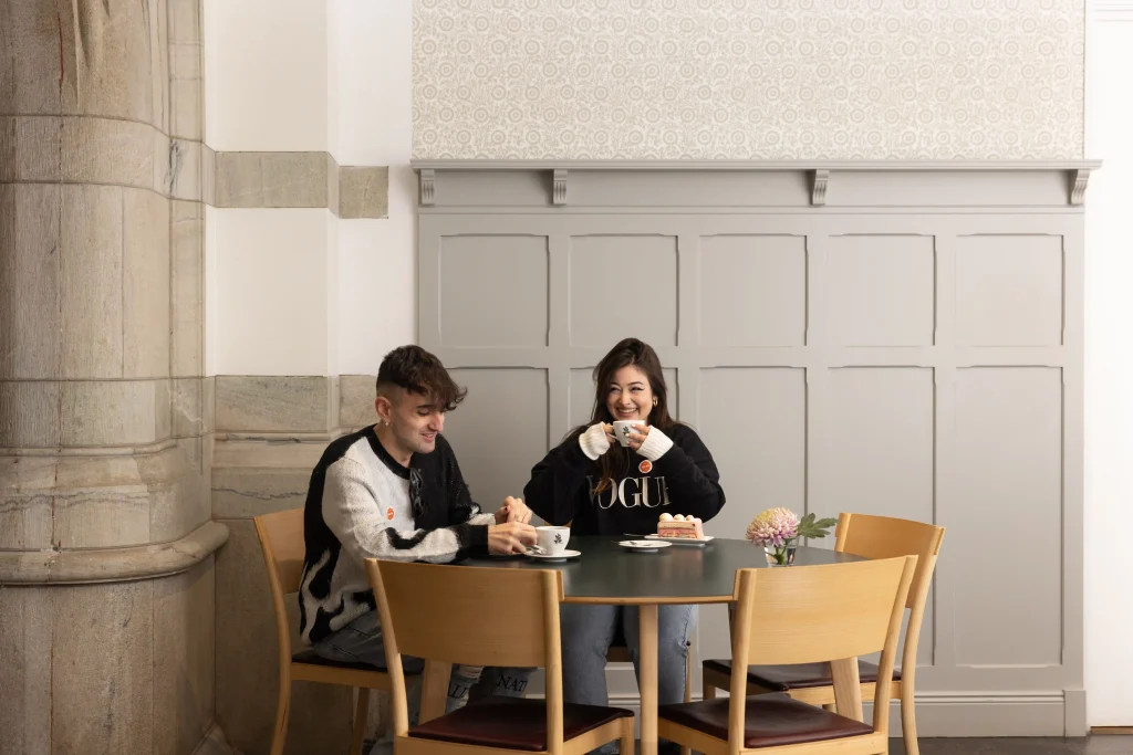 Ett ungt par sitter och skrattar och dricker en cappuccino vid ett bord i en ljus restaurang med grå träpanel, mönstrad tapet och marmorpelare.