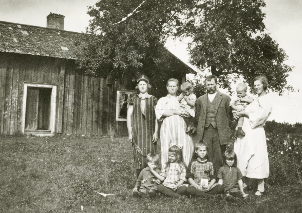 En statarfamilj med tio barn i lolika åldrar står uppställda utanför ett torp, fyra barn sitter i gräset, ett träd i bakgrunden.