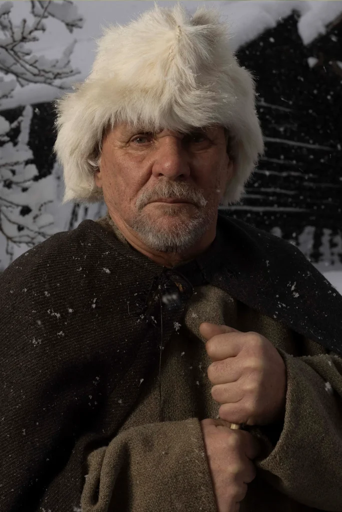 En äldre skäggig man med vit pälsmössa och ylleklädnad står i ett snölandskap med timrade stugor bakom sig.