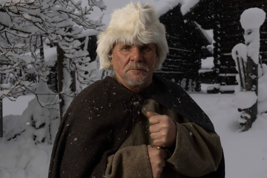En äldre skäggig man med vit pälsmössa och ylleklädnad står i ett snölandskap med timrade stugor bakom sig.