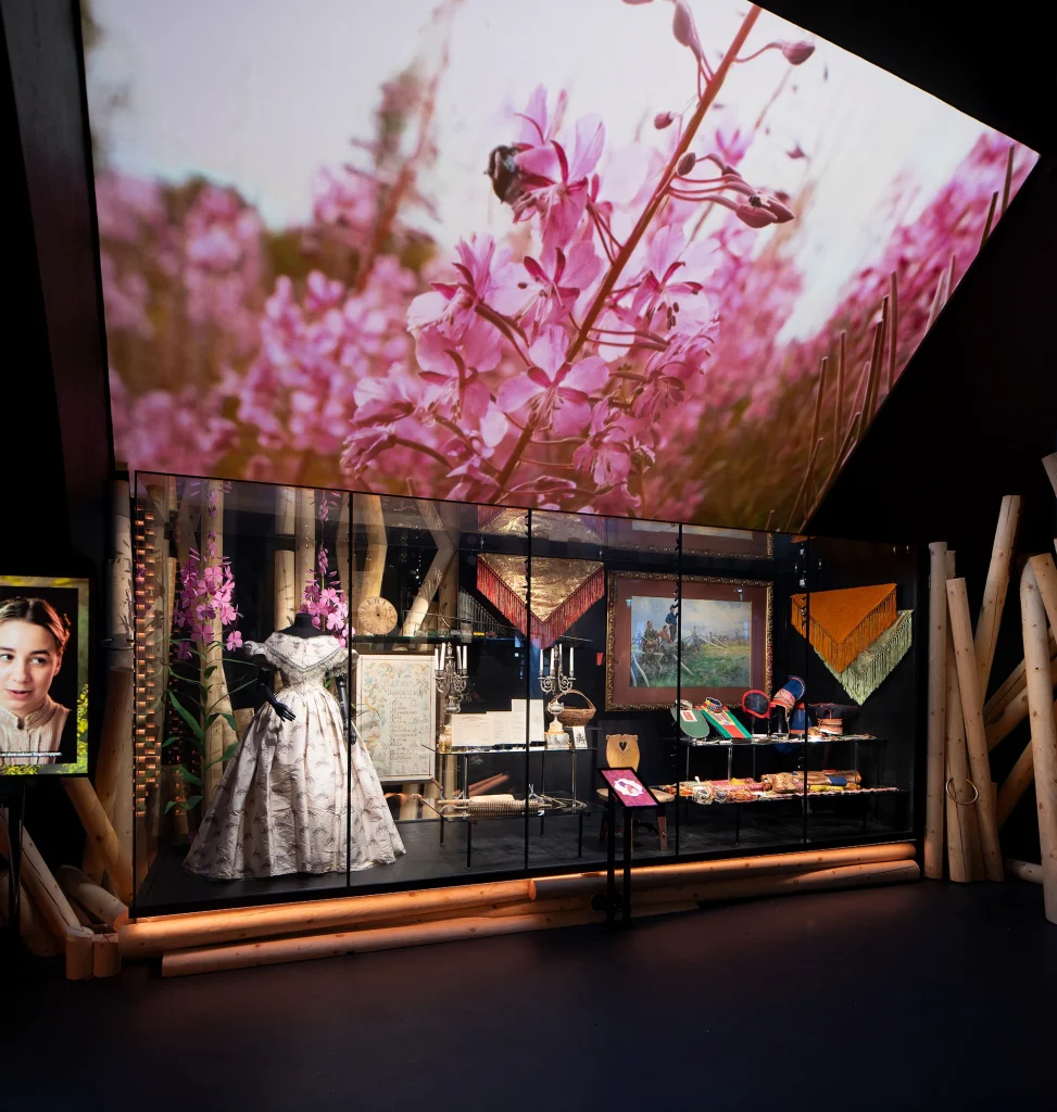 En vid ljust blommig sidenklänning och flera föremål i en stor monter, ovanför en filmduk som visar insekter som pollinerar rosa blommor, till vänster om montern en filmskärm med en ung flicka.