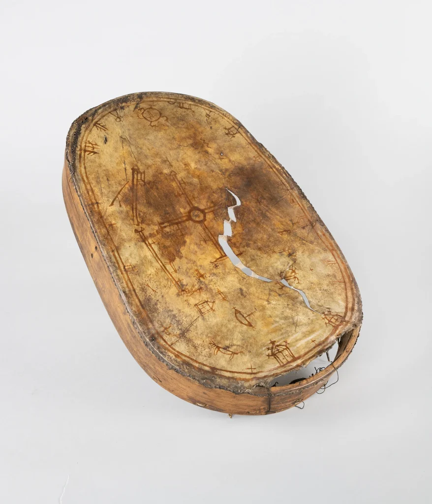En samisk trumma i form av en oval träram med ett skinn spänt runtom, på skinnet finns symboler och tecken, skinnet är gulbrunt och träramen brun.